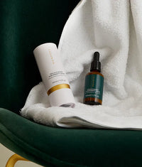 Follicle Boost Hair Density Serum in amber glass bottle with dropper and white cylindrical packaging on white towel and green chair