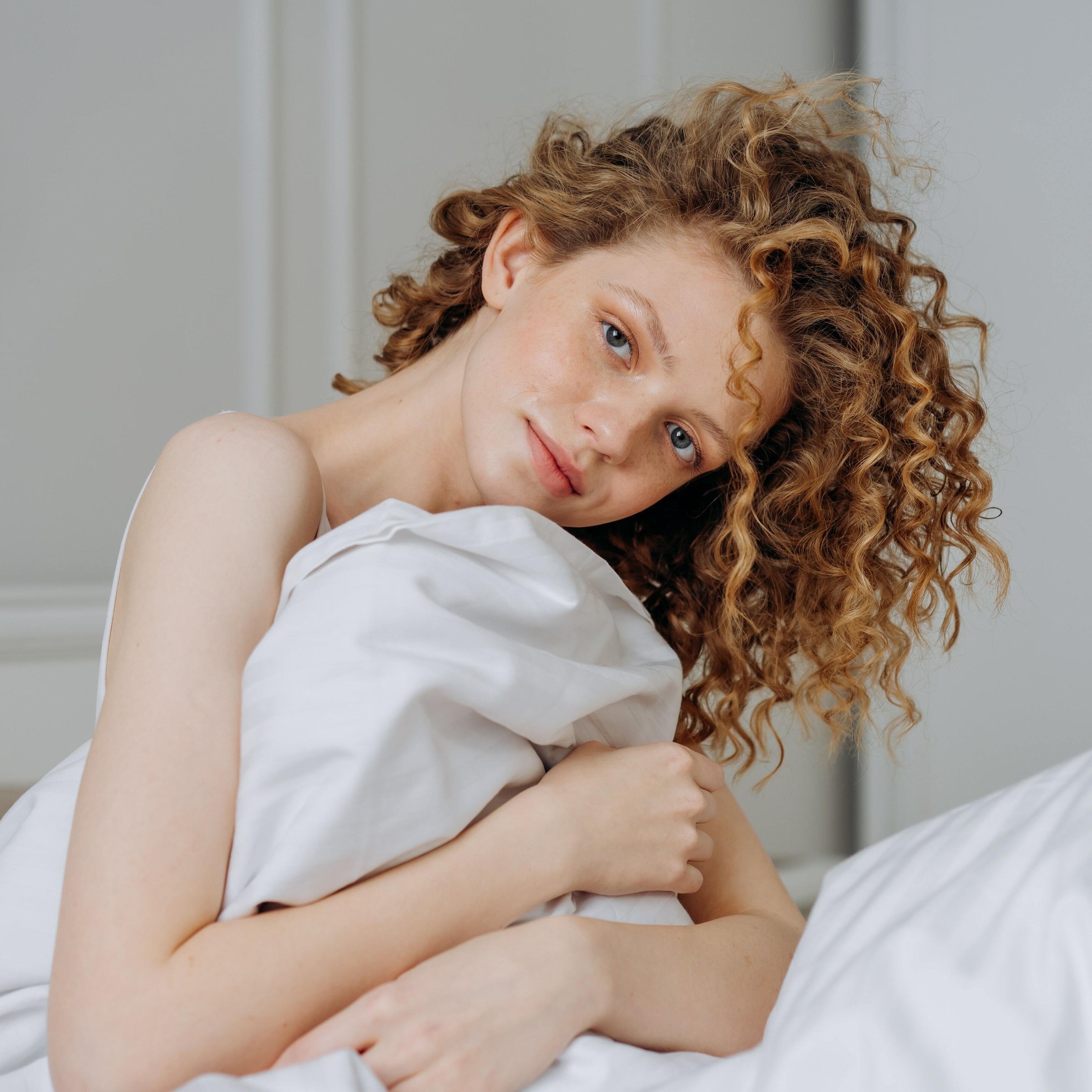 Young woman with voluminous curly auburn hair hugging white bedding in softly lit bedroom