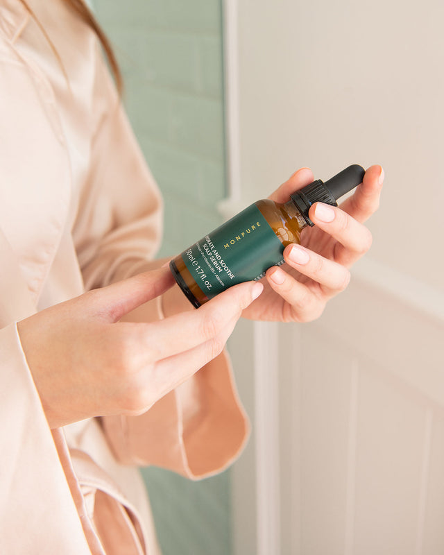 Hands holding amber glass bottle with black dropper and green label for scalp serum