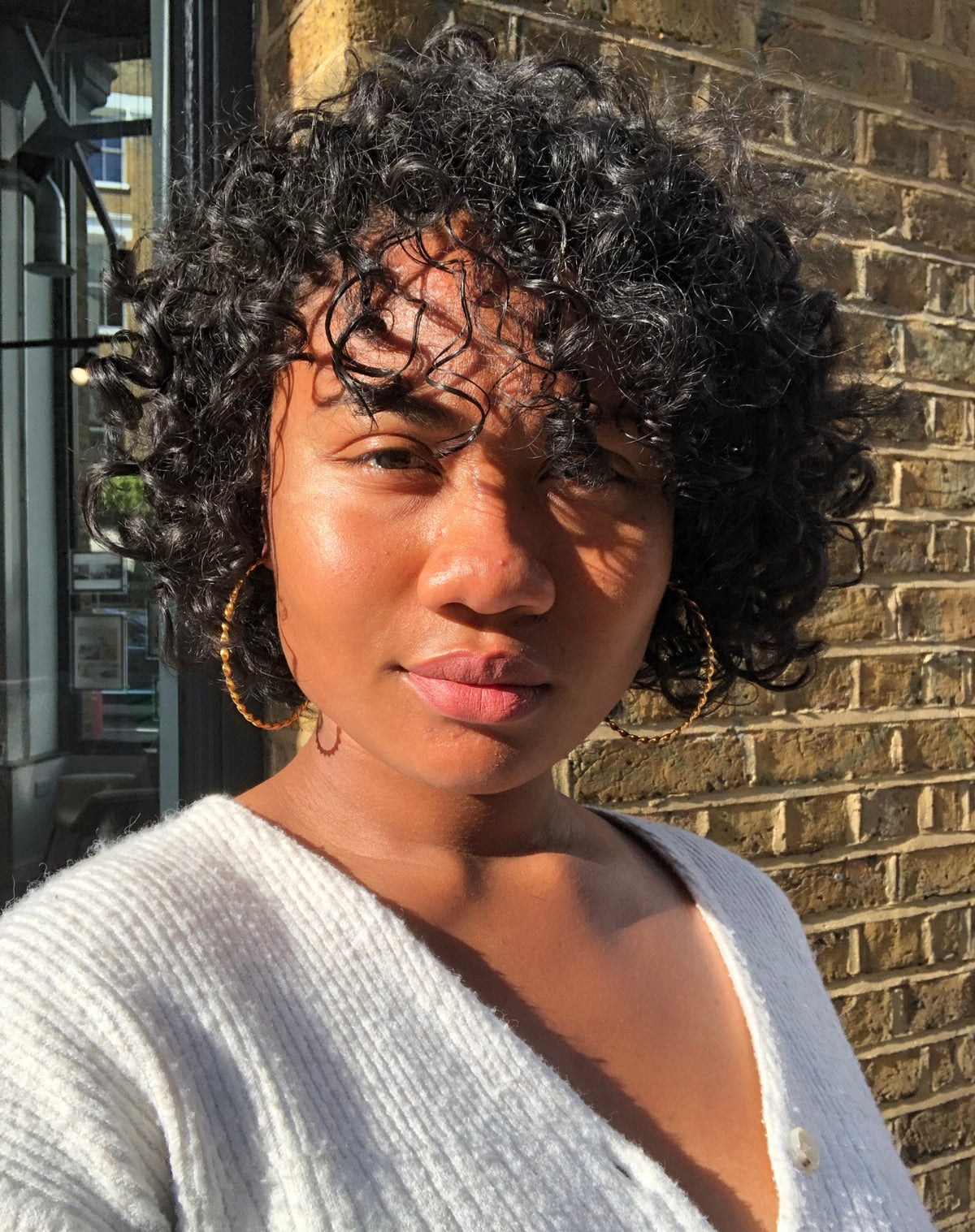 Woman with short natural curly black hair wearing gold hoop earrings and a white knit cardigan
