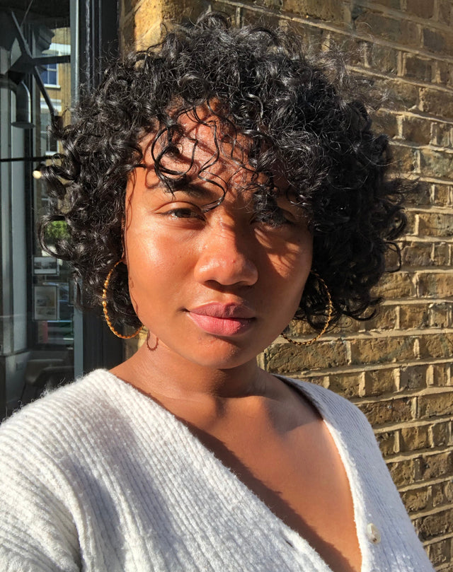 Woman with short natural curly black hair wearing gold hoop earrings and a white knit cardigan