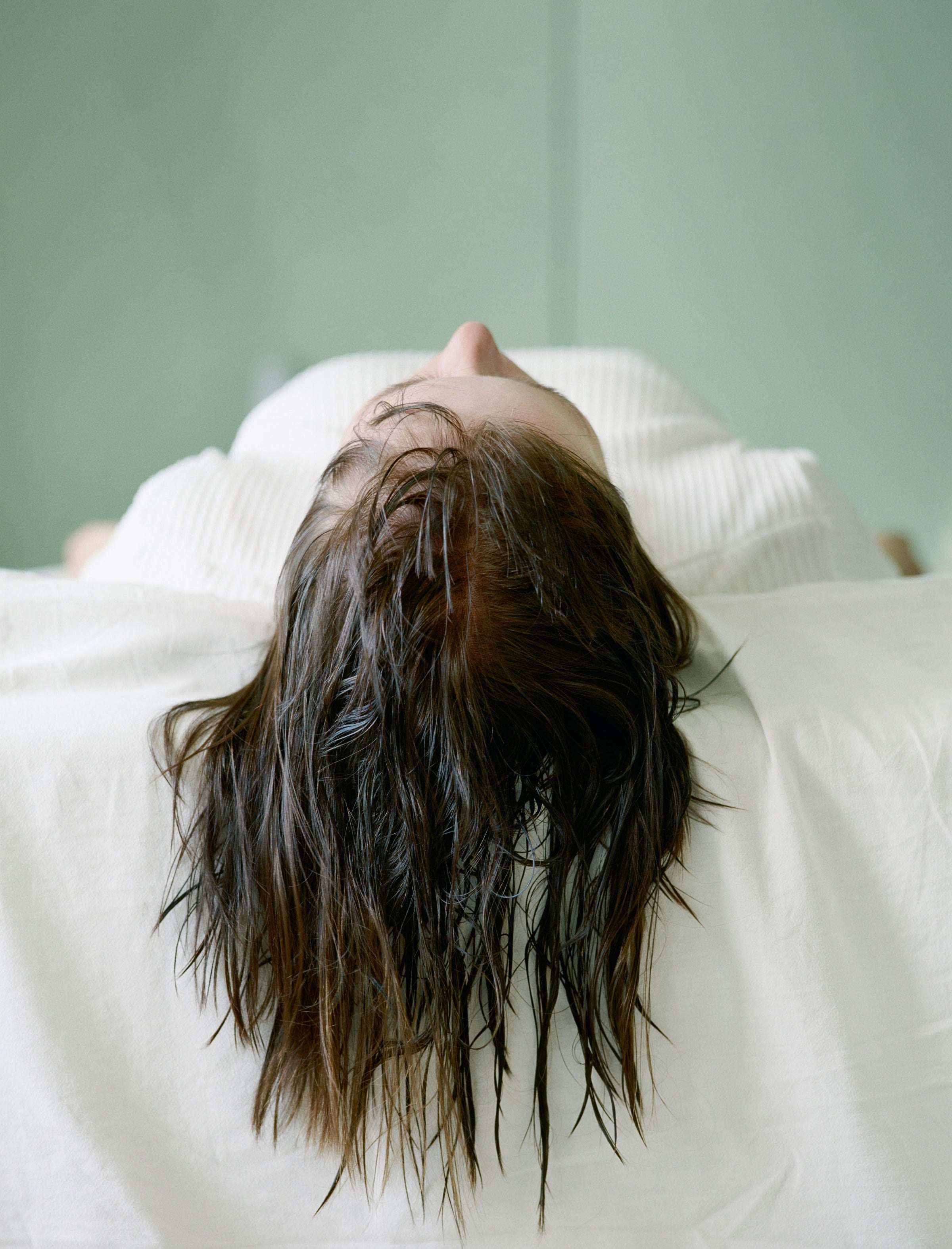 Person lying back with wet, oily brown hair hanging over white surface against pale green background