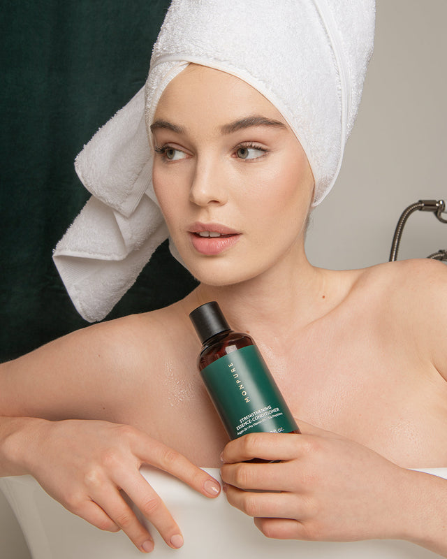 Woman with white towel on head holding green bottle of Monpure Strengthening Essence Conditioner in bath