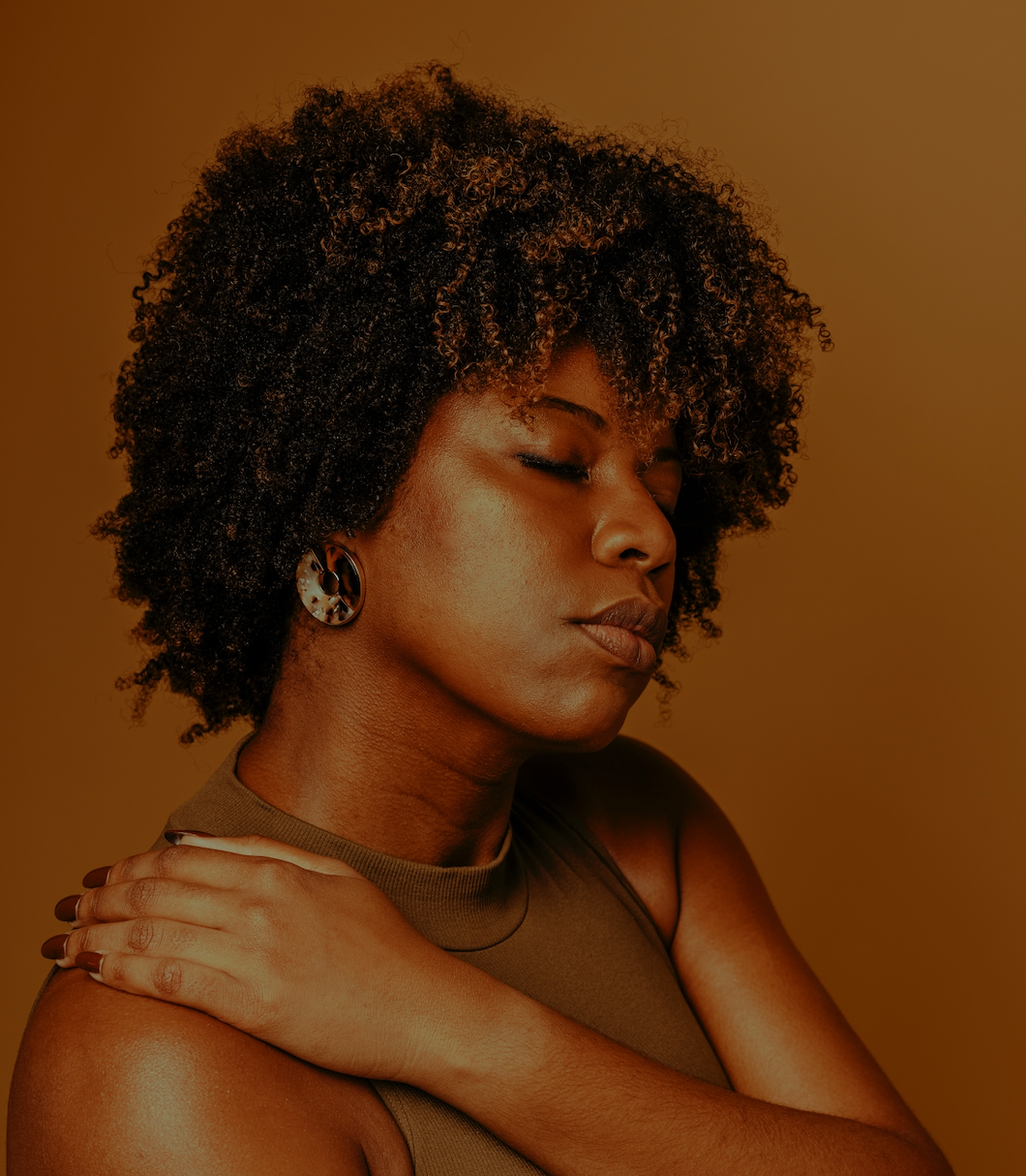 Woman with natural curly hair wearing a sleeveless olive green top and round tortoiseshell earrings