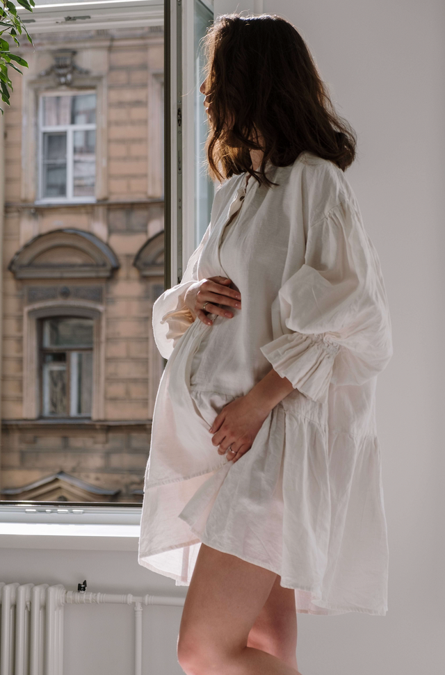 Pregnant woman in loose, white, long-sleeved dress standing by window indoors