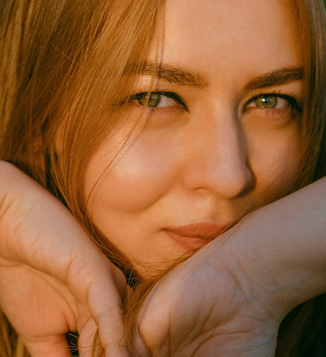 Close-up of a woman with clear skin and light brown hair, gently resting her face on her hands