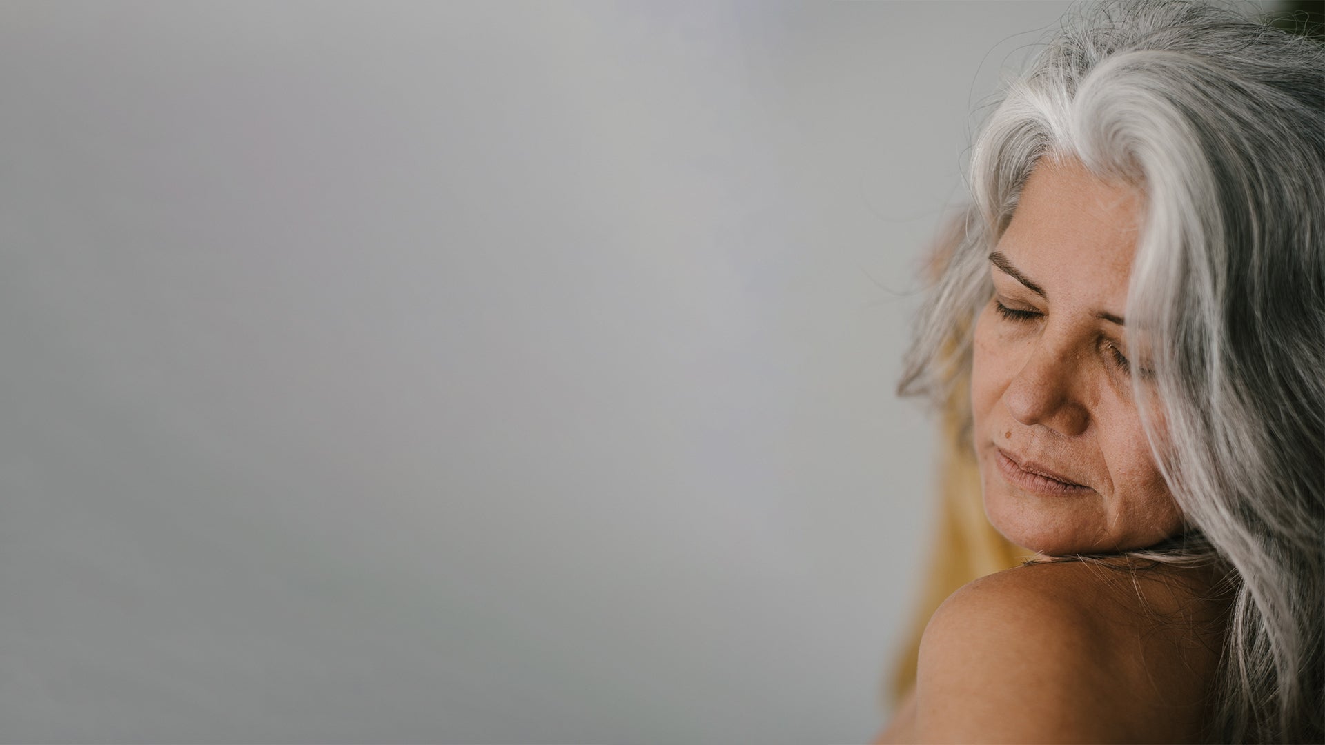 Mature woman with long grey hair and closed eyes against plain grey background