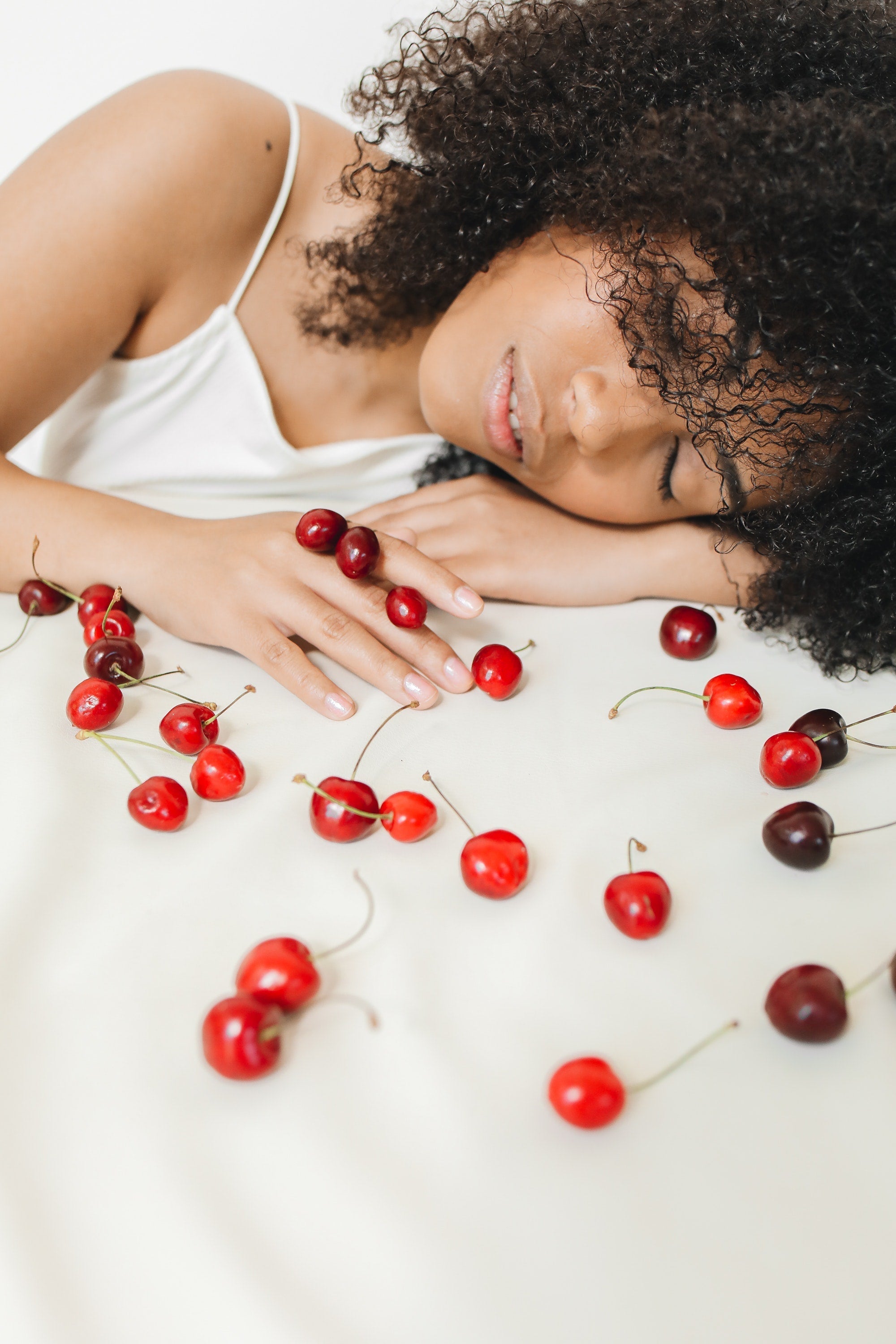 Woman with curly hair resting on white surface surrounded by scattered red cherries