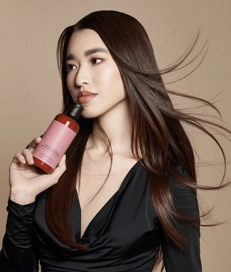 Woman with long dark hair holding a brown bottle with pink label Ceramide Complex Bond Repair Conditioner