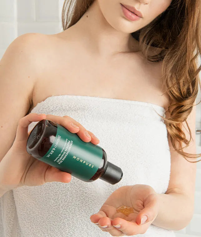 Woman in white towel pouring amber strengthening shampoo from green bottle into hand