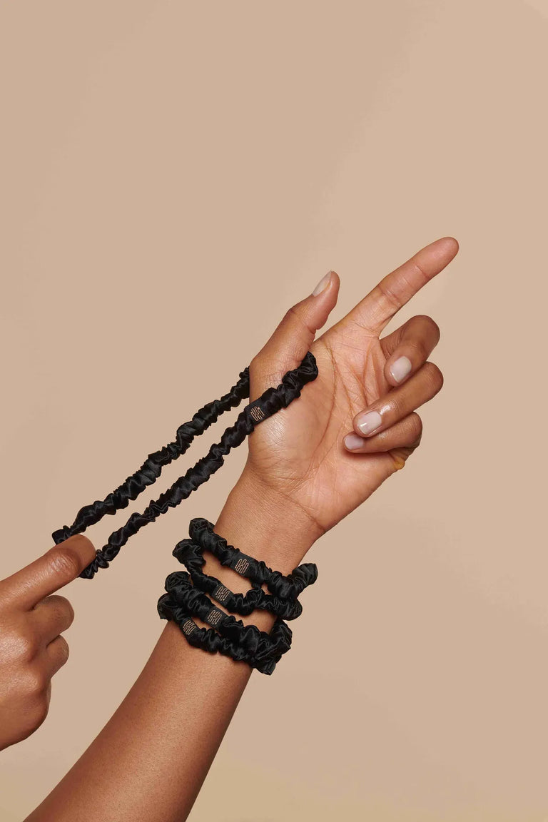 Set of slim black silk scrunchies worn and held by hand against beige background
